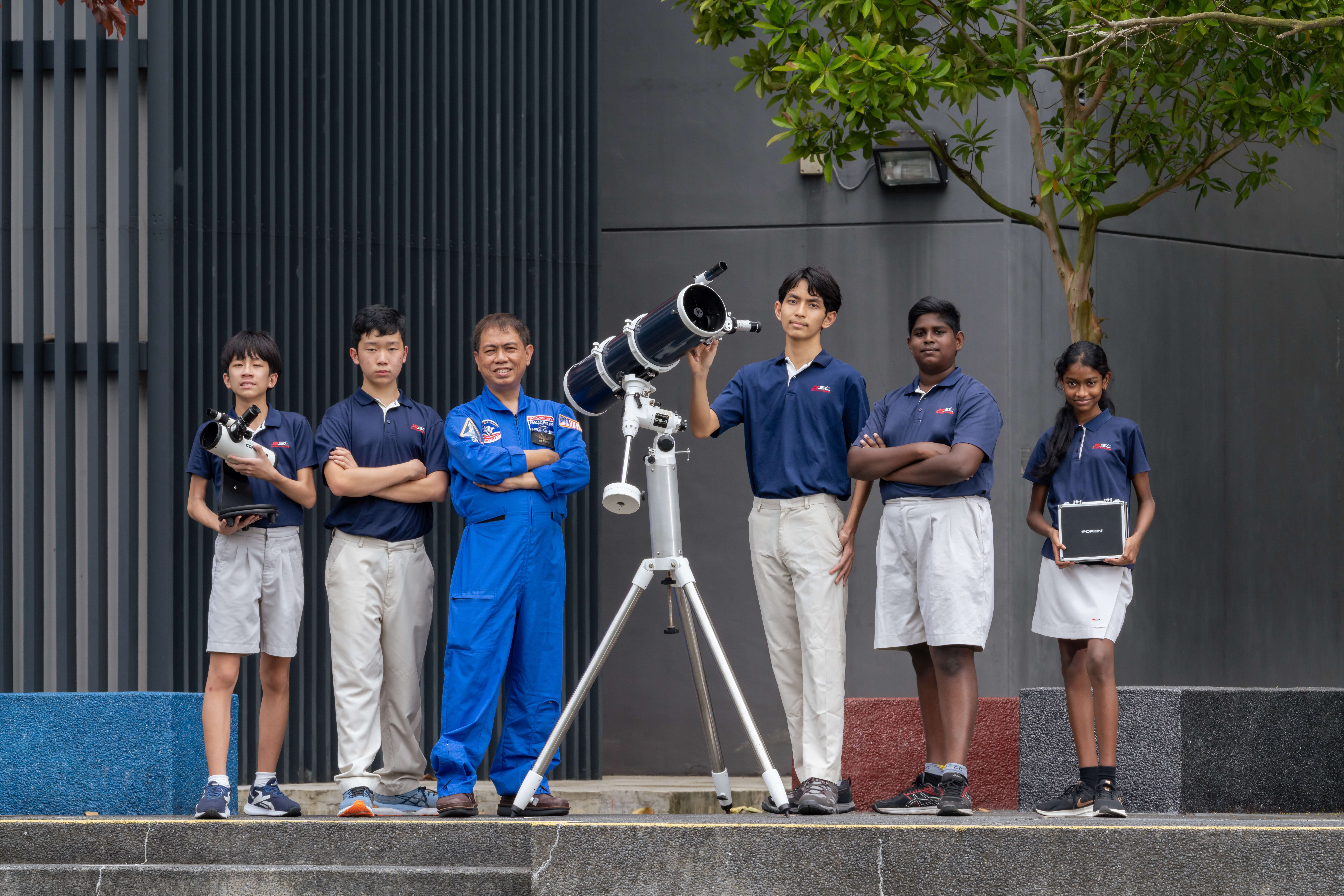 Five students and an adult stand by a telescope on tripod outside a dark wall with a tree overhead.